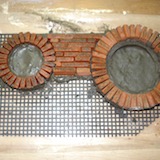 The frontage was built onto a plastic mesh backing. The mesh binds everything together so that the finished item is able to withstand a lot of handling. The bricks were cut from a roofing tile, tumbled to knock the sharp edges off and cemented around a round plastic former.